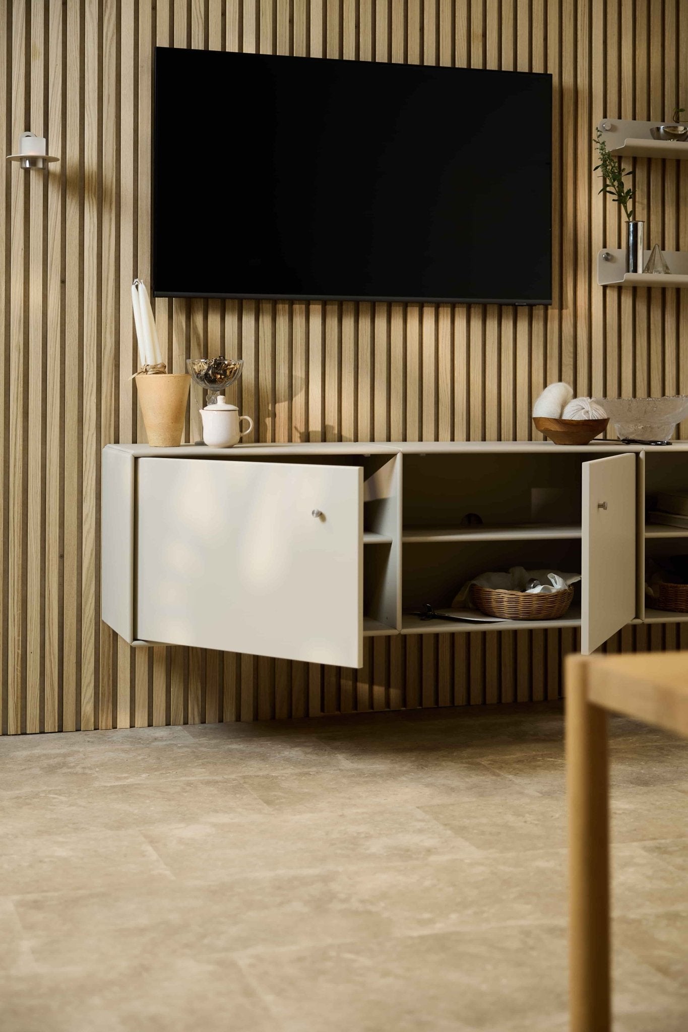 Salón moderno en el que destaca el mueble de televisión Arka en color beige de WoodUpp, situado debajo de un armario y del televisor, sobre una pared acústica nórdica de listones de madera Akupanel. Mobiliario modular.